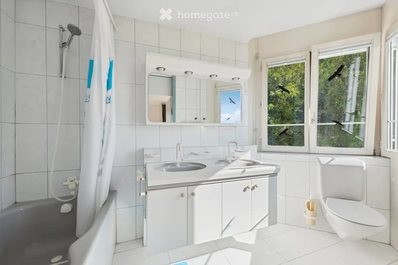 Modern bathroom with white tiles, a bathtub, shower, double sink, mirror, and toilet. Large windows offer a view of greenery outside.