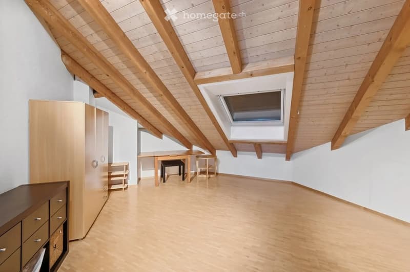 Bright attic room with wooden beams, skylight window, and light wooden flooring. Contains a wardrobe, desk, and chairs.