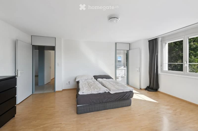 Bright bedroom with wooden flooring, a double bed with white and grey bedding, a nightstand, and large windows with dark curtains. A mirror and an open door lead to a bathroom.