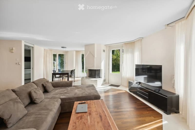 Modern living room with a large sectional sofa, wooden coffee table, TV stand with a flat-screen TV, dining area with a table and chairs, and a fireplace.