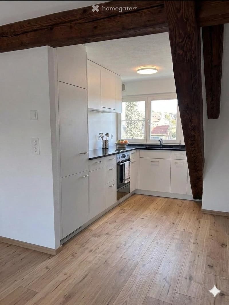 Modern kitchen with white cabinets, wooden flooring, and stainless steel appliances. Features a window with a view of greenery, a bowl on the counter, and a few utensils.