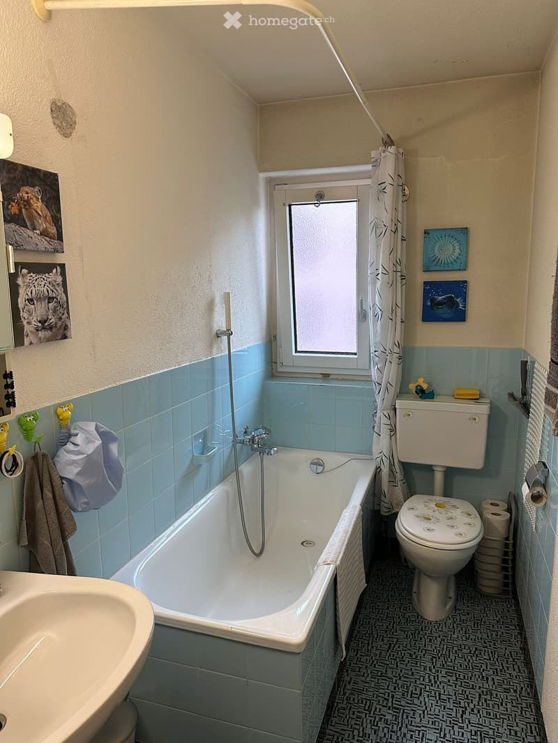Bathroom with a bathtub, toilet, sink, and a window with a shower curtain. Blue tiles on walls and a patterned floor.