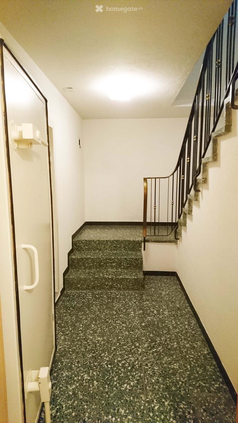 A narrow hallway with a staircase leading upwards, featuring a dark green speckled tile floor and white walls.