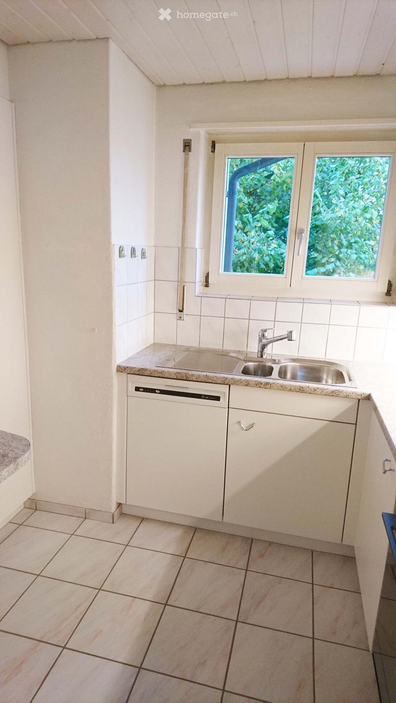 Modern kitchen with white cabinets, granite countertop, stainless steel sink, and dishwasher. Features tiled flooring and large windows with a view of greenery.