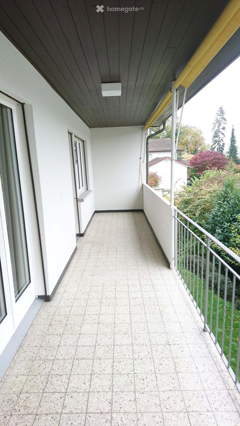 Balcony with tiled floor, white walls, metal railing, and a view of greenery and neighboring buildings.