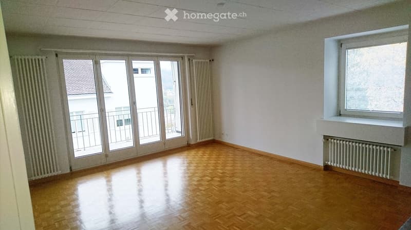 Empty room with wooden flooring, white walls, large sliding glass doors leading to a balcony, and a window with blinds.