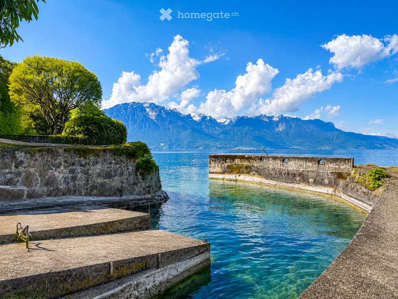 A serene lakeside scene with clear blue water, stone walls, and lush greenery, with snow-capped mountains in the background.