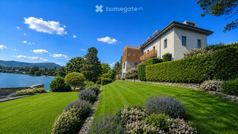 A well-maintained garden with a lawn, flower beds, and shrubs leading to a modern house with a balcony overlooking a lake.
