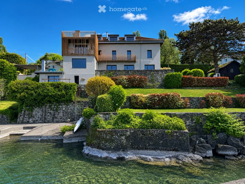 A modern two-story house with a wooden balcony overlooking a serene lake, surrounded by well-maintained gardens and lush greenery.