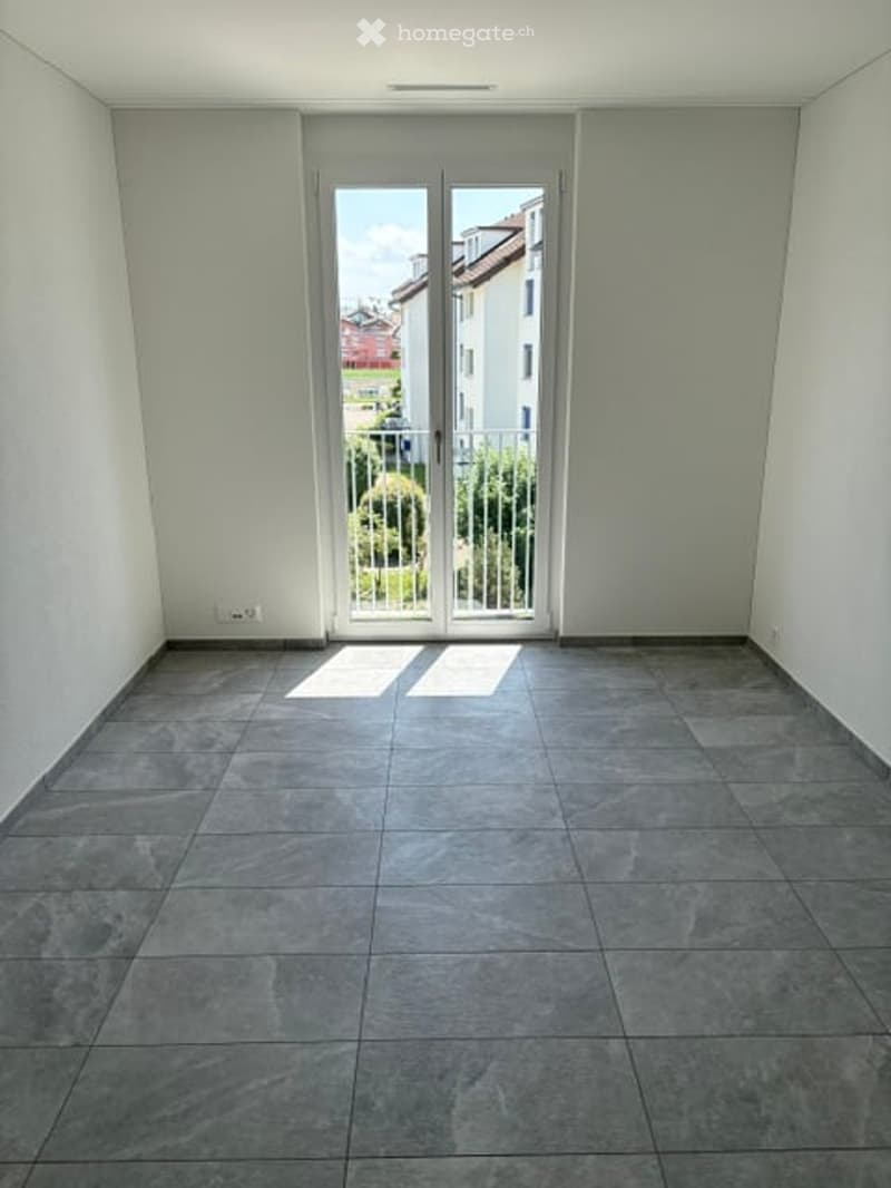 Empty room with grey tiled floor, white walls, and large glass sliding doors leading to a balcony with a view of neighboring buildings and greenery.
