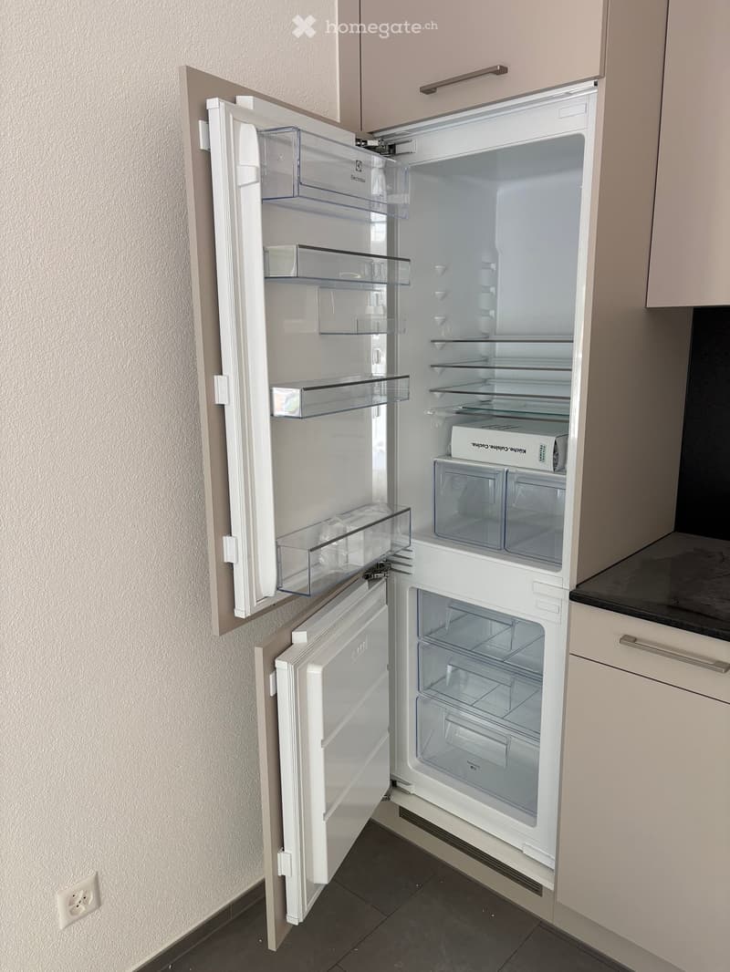 A modern white refrigerator with multiple compartments and shelves, integrated into a kitchen with light-colored cabinets and dark countertops.