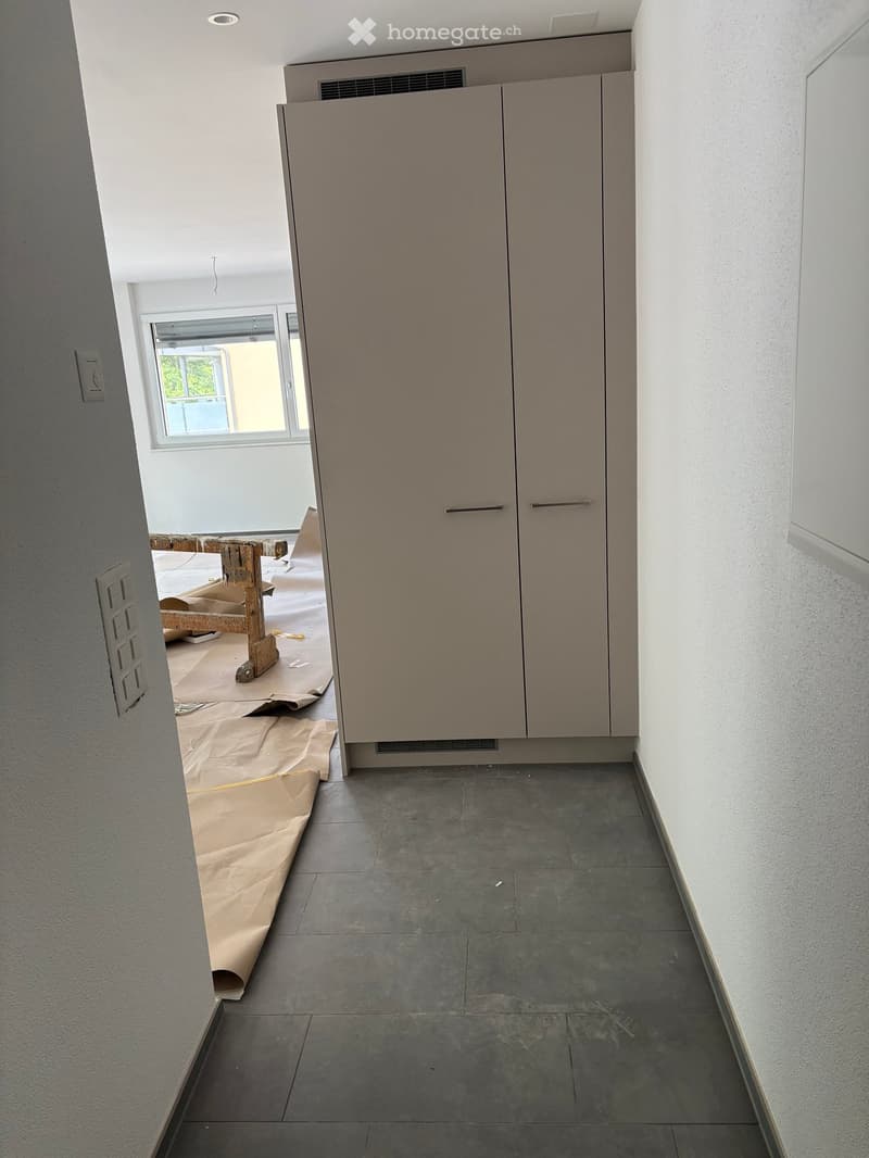 Empty room with grey tiled floor, white walls, and built-in cabinets. The room is under construction with protective coverings on the floor and a partially visible workspace in the background.
