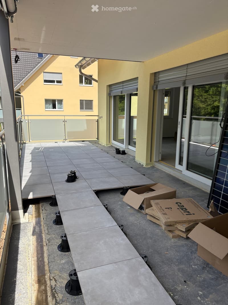 Empty balcony with tiled floor, metal railing, and several black circular objects on the ground, with cardboard boxes stacked on the right side.