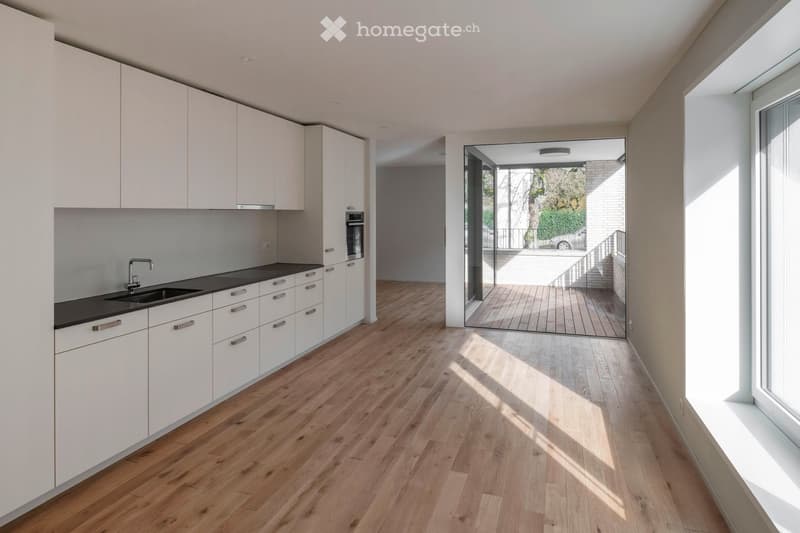 Modern kitchen with white cabinets, wooden flooring, and a black countertop. Features a sink, faucet, and built-in oven. Large window and sliding glass door lead to an outdoor area.
