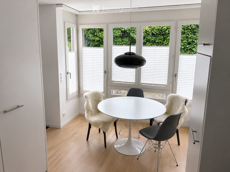 Bright, modern dining area with a white round table, four chairs, and large windows with blinds overlooking greenery.