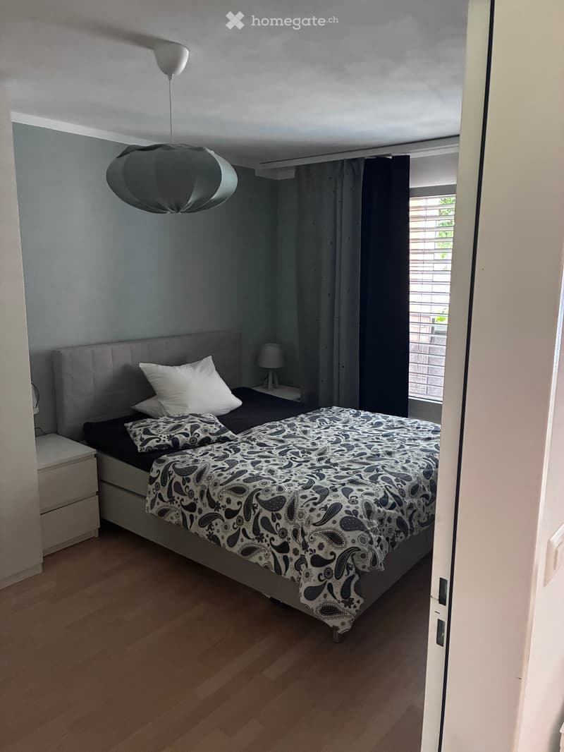 Modern bedroom with a double bed, patterned bedspread, nightstand, and lamp. Wooden flooring and grey walls.