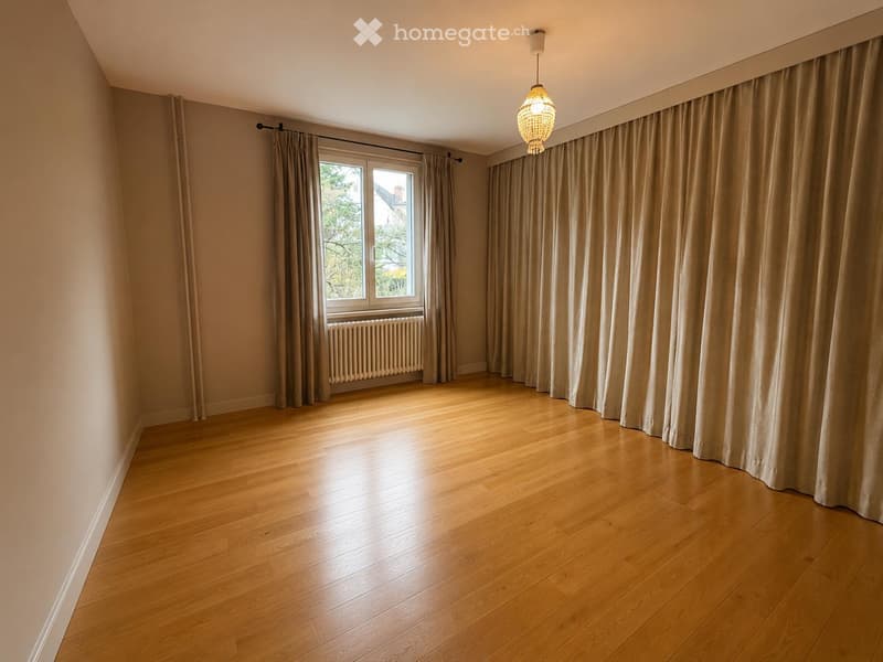 Empty room with wooden flooring, large windows with curtains, radiator, and a hanging light fixture.