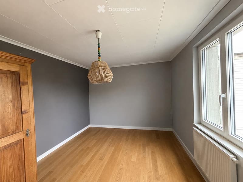 Empty room with wooden flooring, grey walls, large windows, and a hanging woven pendant light fixture.