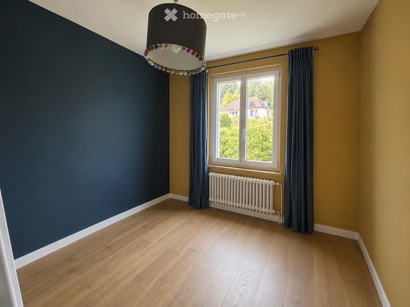Empty room with wooden flooring, dark blue and yellow walls, large window with curtains, radiator, and a hanging lamp with colorful accents.