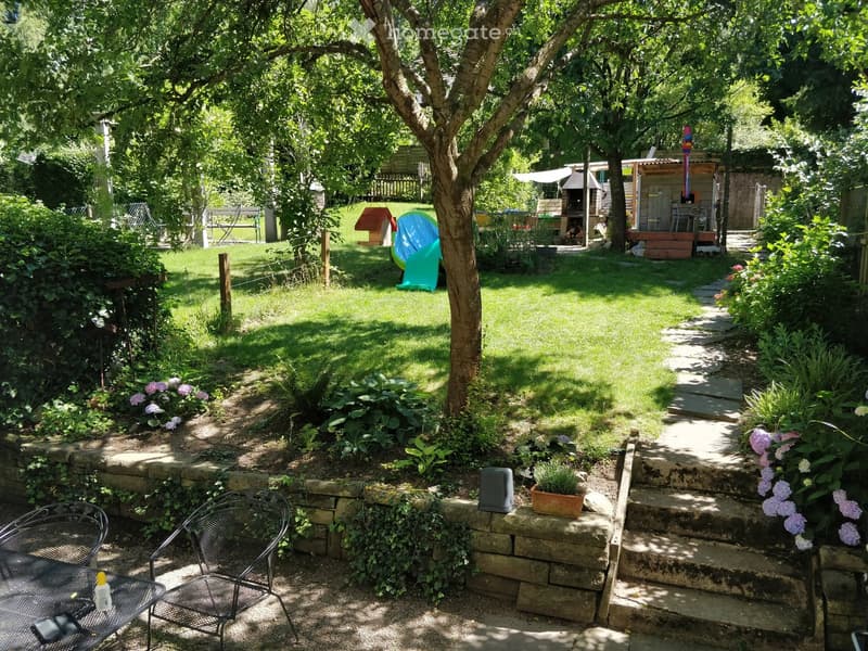 A well-maintained garden with a stone pathway, steps, a small seating area with a table and chairs, and a potted plant. The garden features lush greenery, flowers, and a small shed in the background.