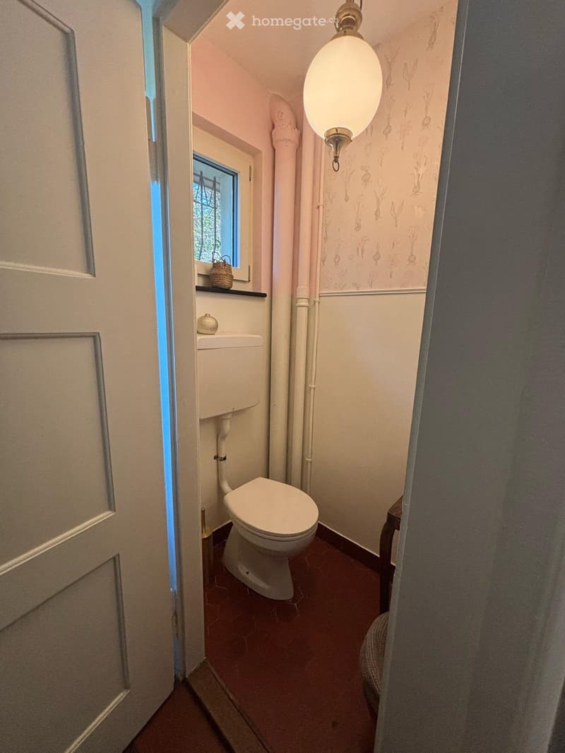 A compact bathroom with a toilet, a small window, a shelf with a basket, and a decorative light fixture.