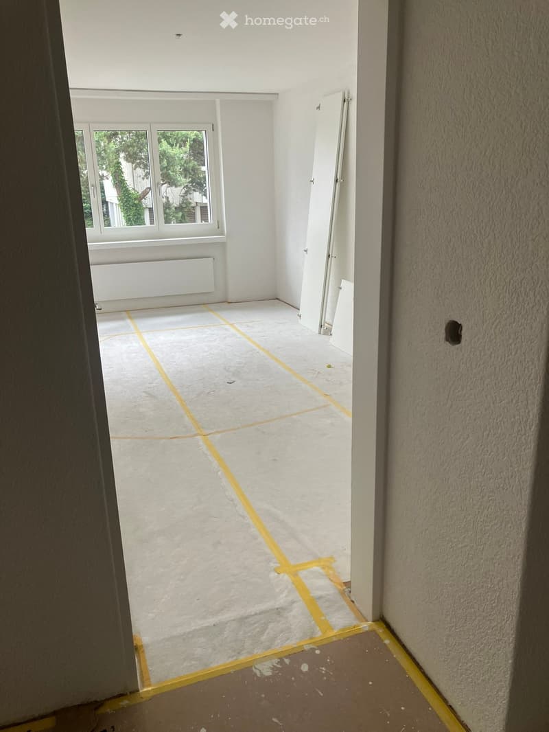 Empty room with white walls, concrete floor, and a window with white frame. Yellow tape marks the floor area.