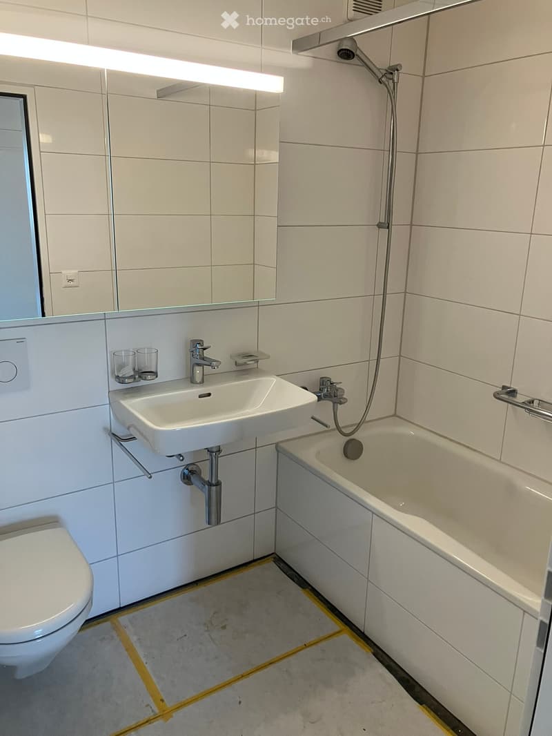 Modern bathroom with white tiles, a toilet, a sink with a faucet, a bathtub, a mirror, and two cups on the sink counter.