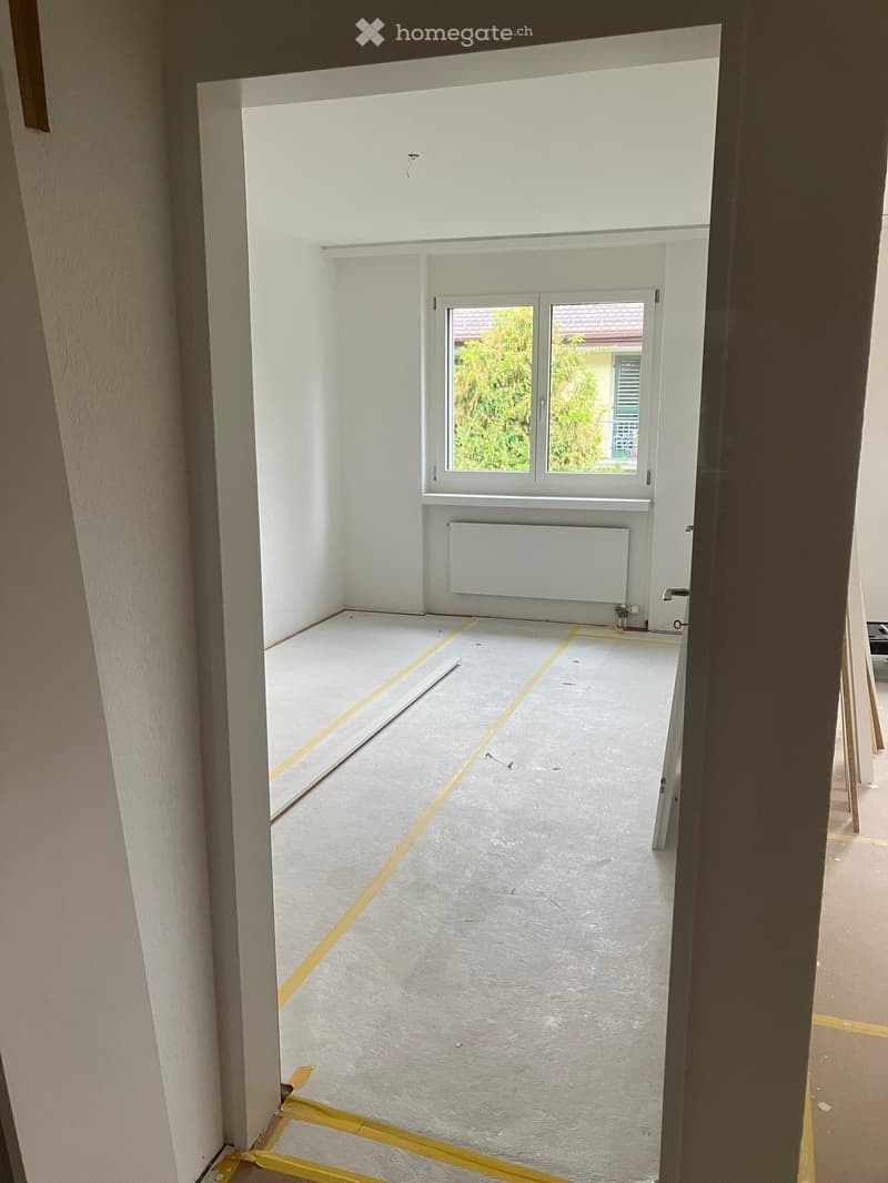 Empty room with white walls, a window, and a radiator. The floor is covered with protective material and yellow tape, indicating ongoing renovation.