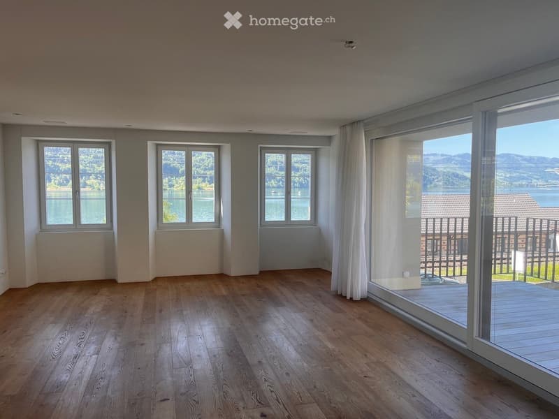 Unfurnished Room. Empty room with wooden flooring, large windows, and a view of a lake and mountains. The room has white walls and a sliding glass door leading to a balcony.