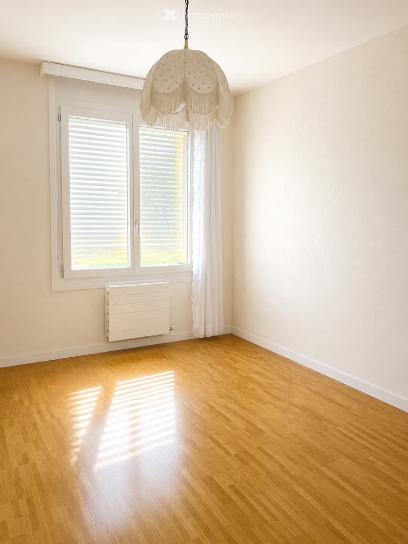 Empty room with wooden flooring, white walls, large window with blinds, radiator, and a decorative hanging light fixture.