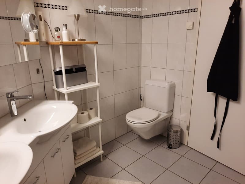 Modern bathroom with white tiles, a sink, toilet, mirror, and a shelf holding towels and toiletries.