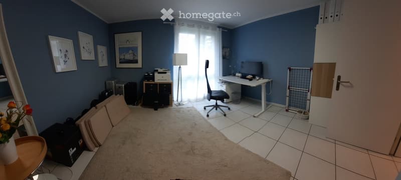 A well-lit room with blue walls, a white tiled floor, and a large window with white curtains. The room features a desk with a chair, a lamp, a laptop, and a printer. There are framed pictures on the wall, a radiator, a small table with a vase of flowers,