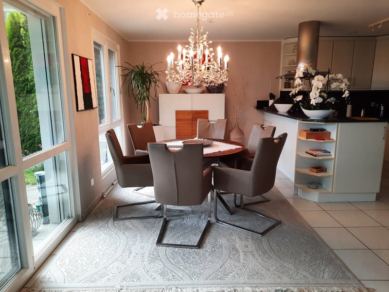 Modern dining area with a round wooden table, six leather chairs, a large chandelier, and a kitchen area with cabinets and shelves. The space features a patterned rug and large windows with a view of greenery.