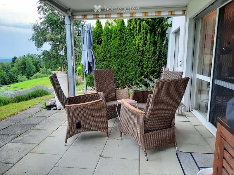 Balcony with wicker chairs and a small table, overlooking a green landscape with trees and a view of the valley.