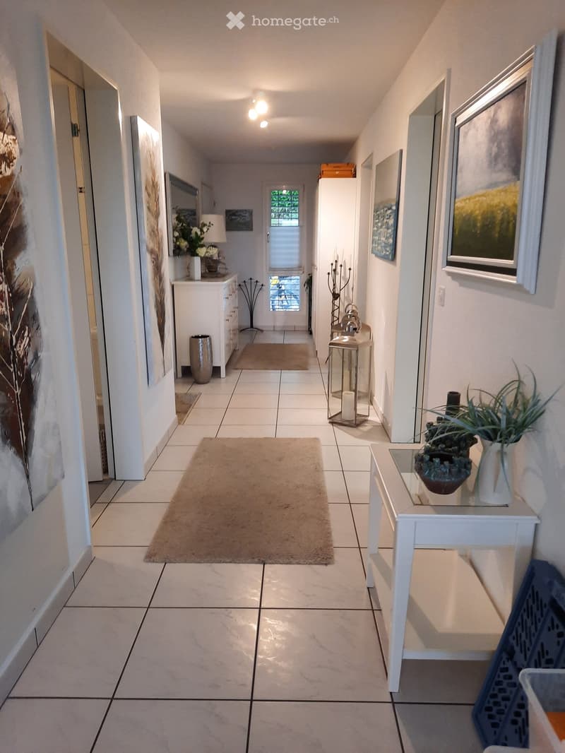 A well-lit hallway with tiled flooring, white walls, and several decorative items including a small table with a plant, a lantern, and artwork on the walls.
