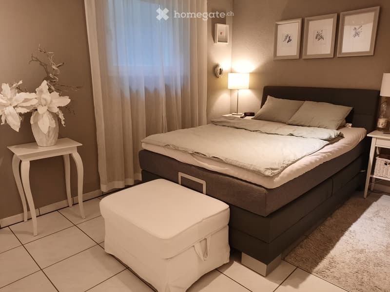 Modern bedroom with a large bed, nightstand, stool, side table with vase, and wall-mounted lamp. Light-colored bedding and neutral walls.