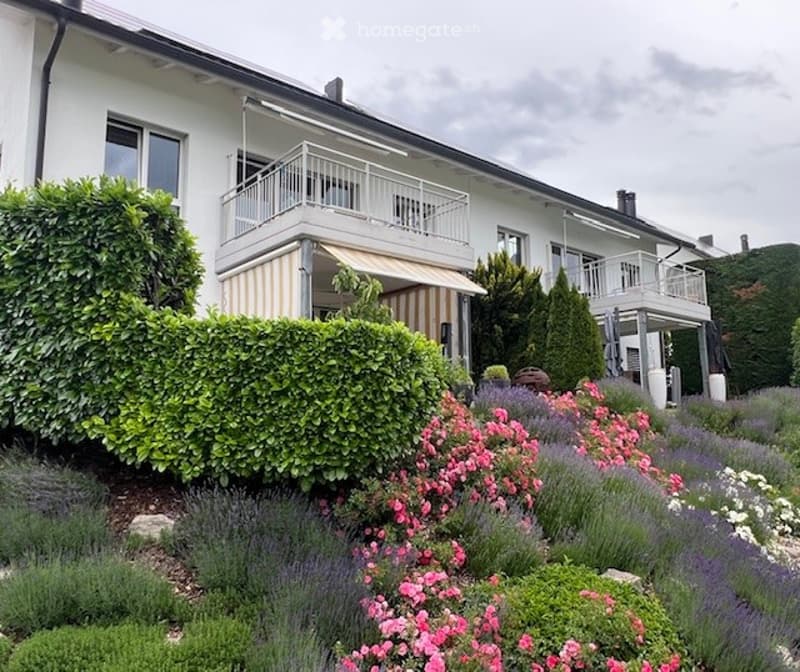 Well-maintained garden with colorful flowers, shrubs, and a neatly trimmed hedge. The house features balconies with white railings and a striped awning.