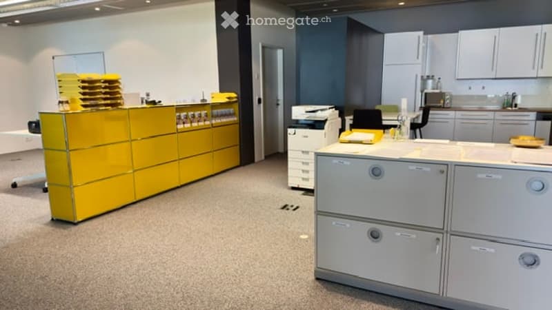 Modern office space with a bright yellow reception desk, grey filing cabinets, a small kitchen area with cabinets and a sink, and a printer on a stand. The room features a clean, organized layout with neutral-colored carpeting and white walls.