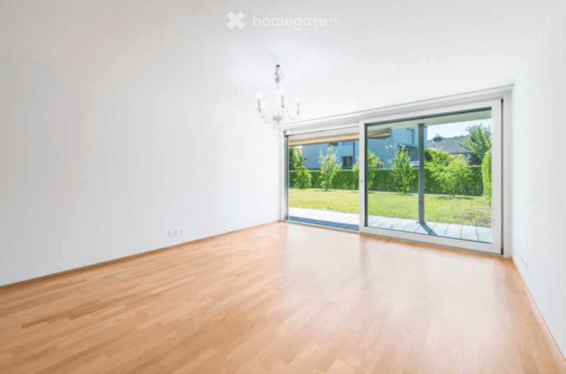 Empty room with light wooden flooring, white walls, and large sliding glass doors leading to a garden area.