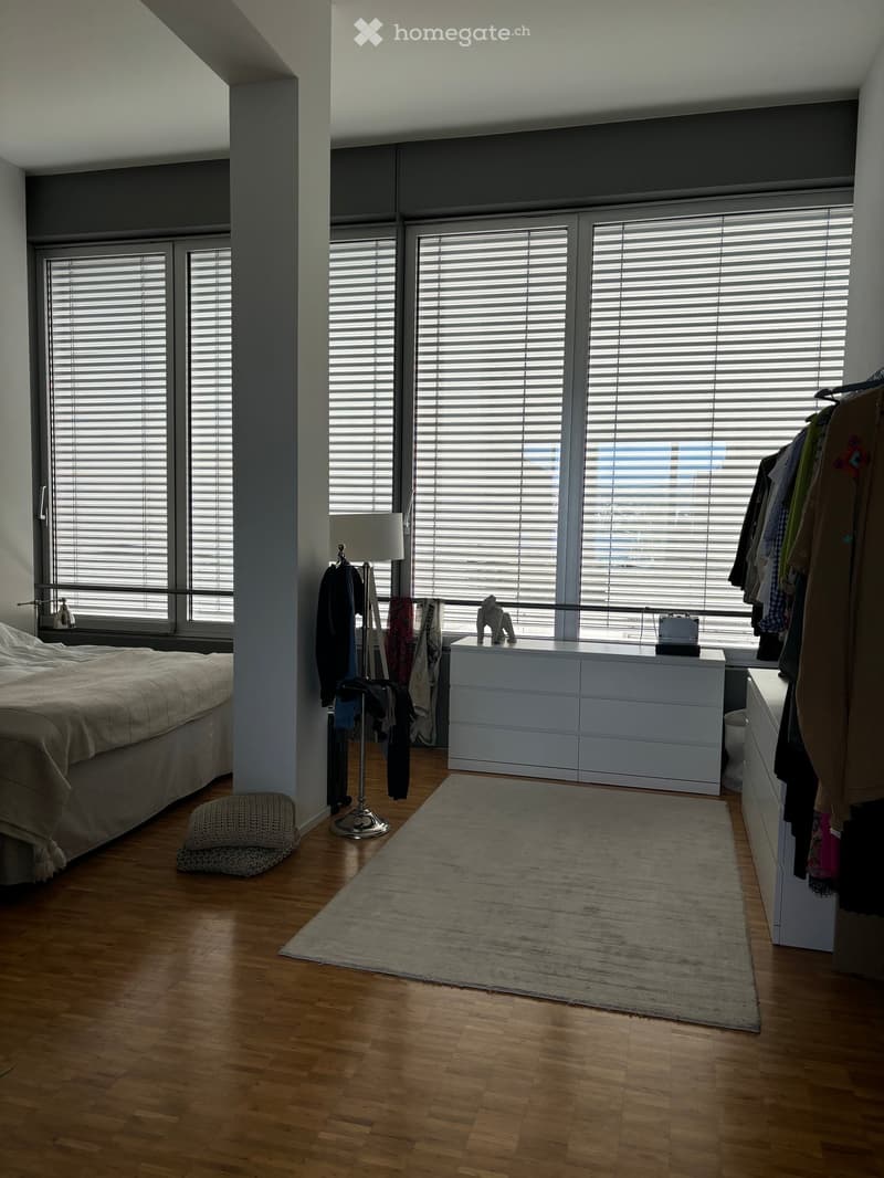 Bright bedroom with wooden flooring, large windows with blinds, a bed with a beige duvet, a nightstand with a lamp, a dresser, a rug, and a clothing rack with hanging clothes.