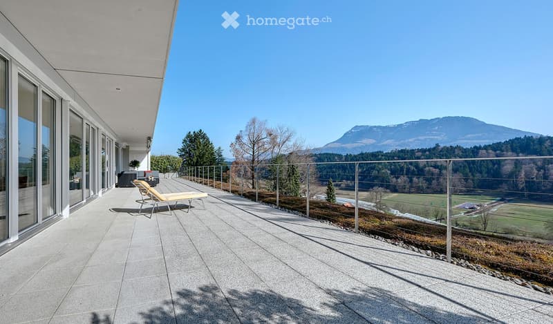 Modern balcony with a panoramic mountain view, featuring a lounge chair, glass railing, and large windows.