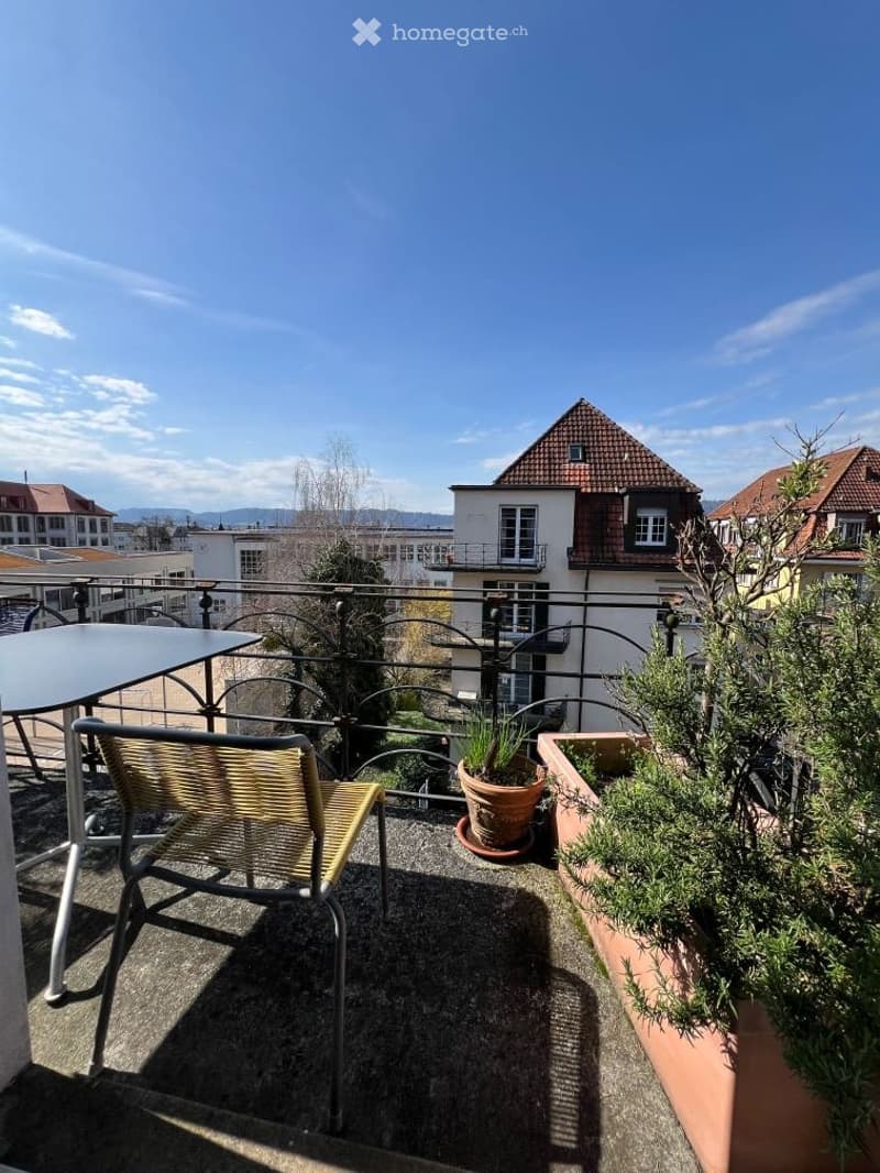 Balcony with a view of the city, featuring a metal railing, a wicker chair, a small table, and potted plants.