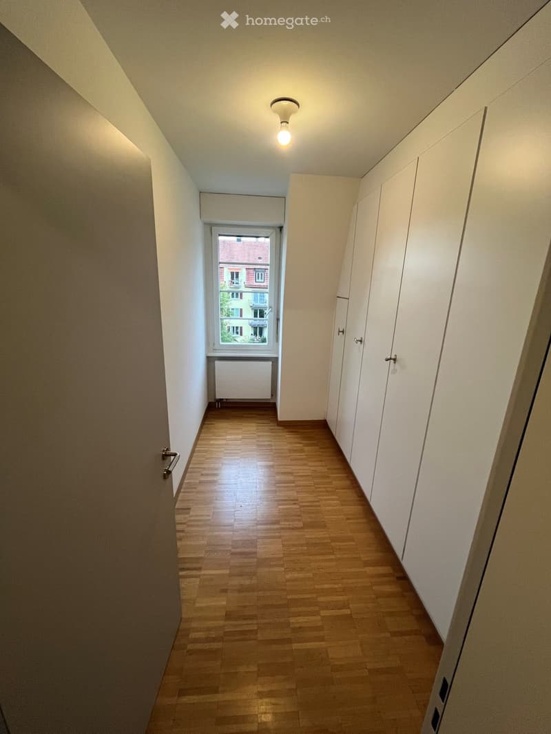 Empty hallway with wooden flooring, white walls, and a window showing a view of a building outside. Features a radiator and built-in storage units.
