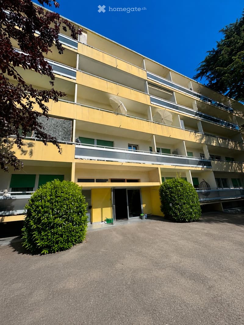Multi-story residential building with yellow and white facade, multiple balconies, and greenery around the entrance.