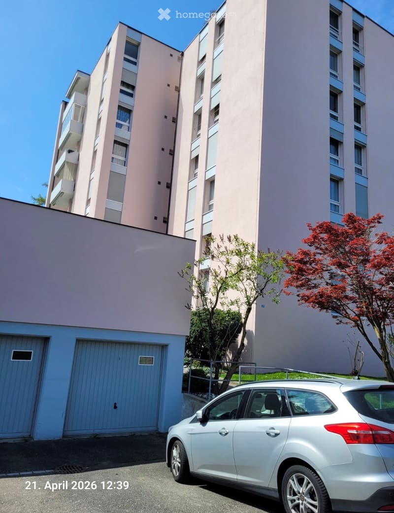 Modern multi-story residential building with light pink facade, balconies, and a garage entrance. A silver car is parked in front.