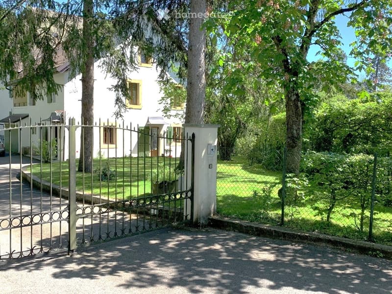 A white two-story house with yellow-framed windows, surrounded by a well-maintained garden with trees and a metal gate entrance.