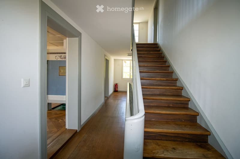 A narrow hallway with wooden flooring and white walls, featuring a staircase with wooden steps and a white handrail leading upwards. A red fire extinguisher is visible near the staircase, and an open doorway leads to another room with blue walls.
