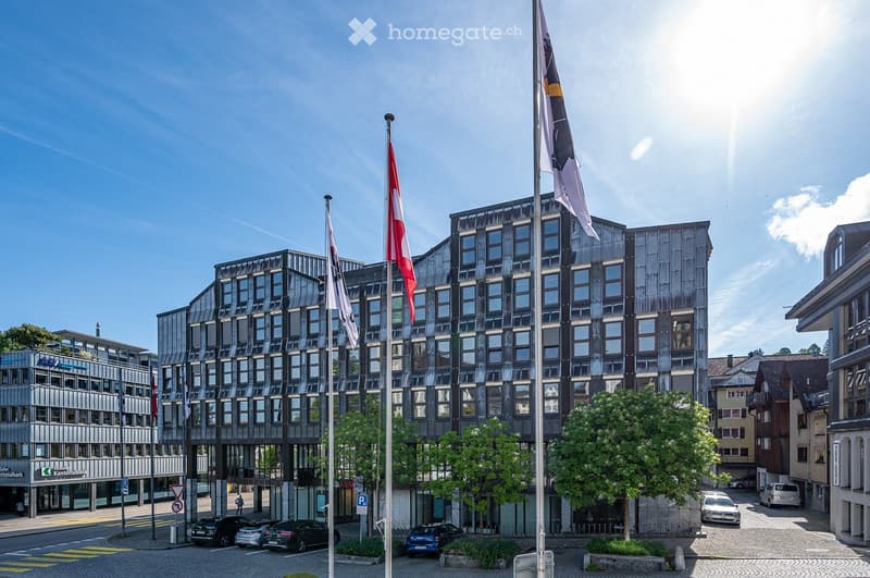 Modern multi-story building with a sleek, dark facade and numerous windows, surrounded by trees and parked cars.