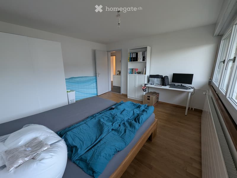A furnished bedroom with a bed, desk, chair, computer, bookshelf, radiator, and backpack. The room has wooden flooring and white walls.