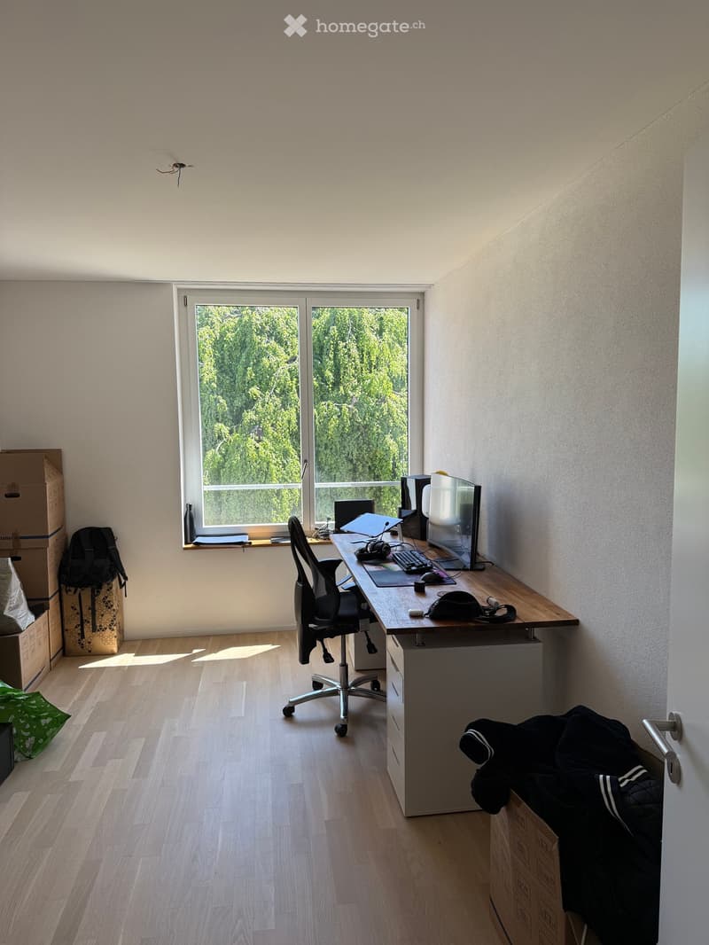 A modern, well-lit room with wooden flooring, a large window offering a view of greenery, a desk with a computer setup, and a comfortable office chair.